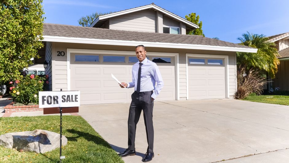A single, professional real estate agent stands confidently with a welcoming smile in front of a beautiful suburban house.